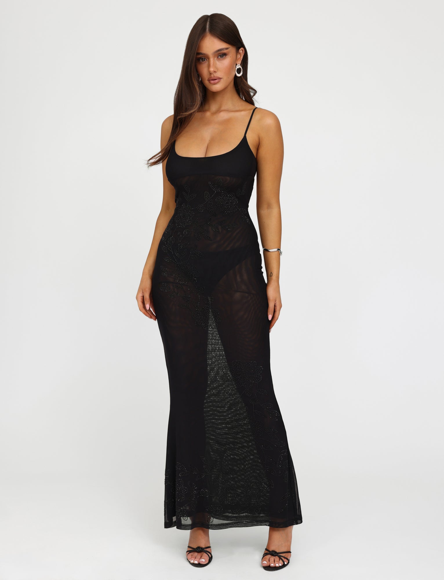 A woman with long hair wears the OCTAVIA DRESS - BLACK from TIGERMIST, a sleeveless, form-fitting, sheer black dress featuring intricate patterns throughout. She pairs the dress with black high-heeled sandals, hoop earrings, and a bracelet on her left wrist. The background is plain white.