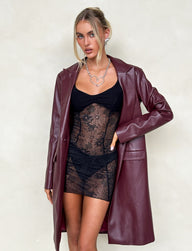 A woman poses against a plain background wearing the LYON TRENCH COAT in cherry red from TIGERMIST over a lacy black mini dress. Her hair is styled in a sleek updo with some loose strands framing her face, and she completes the look with a silver heart-shaped necklace and minimal makeup.