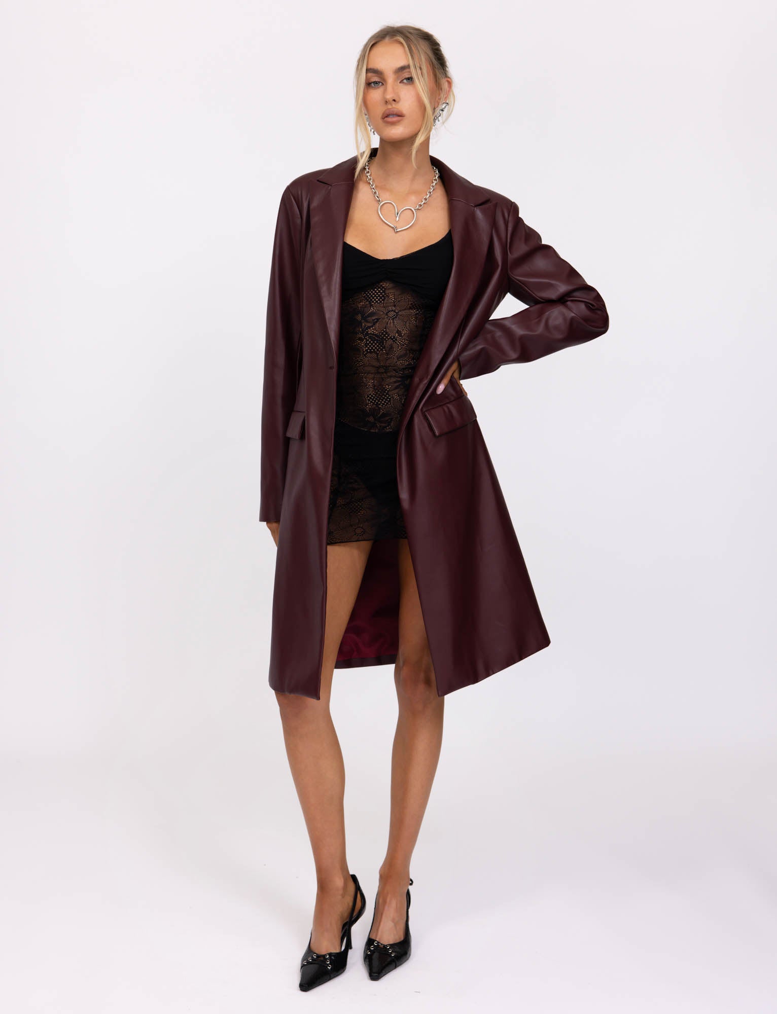 A person with blonde hair in an updo wears the Cherry Red Lyon Trench Coat from TigerMist over a black lace dress. They stand with one hand holding the coat open and the other arm down by their side, also wearing black pointed-toe heels and a statement necklace.