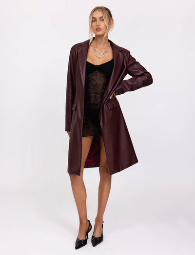 A person with blonde hair in an updo wears the Cherry Red Lyon Trench Coat from TigerMist over a black lace dress. They stand with one hand holding the coat open and the other arm down by their side, also wearing black pointed-toe heels and a statement necklace.