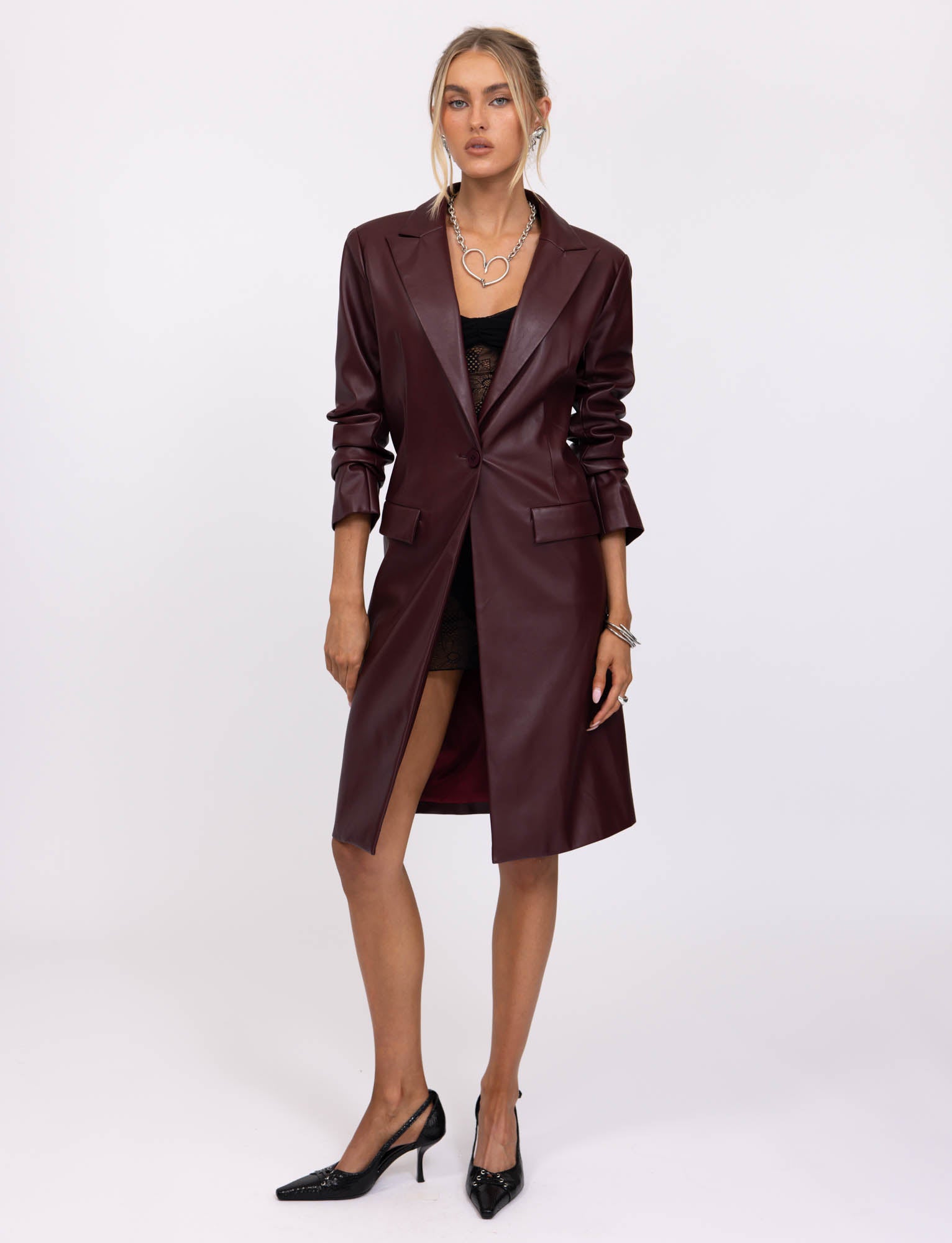 A person with long hair stands against a plain background wearing a cherry red Lyon Trench Coat from TIGERMIST over a black lace dress. They have black pointed-toe heels and accessorize with a silver necklace and bracelet. They have a relaxed yet confident posture.