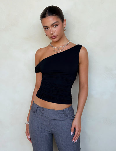 A person with neatly tied-back dark hair wears the stylish TIGERMIST KADENCE TOP in black and gray pants, complemented by silver jewelry including a necklace and earrings. They stand against a light-colored textured wall, gazing directly at the camera with a confident expression.
