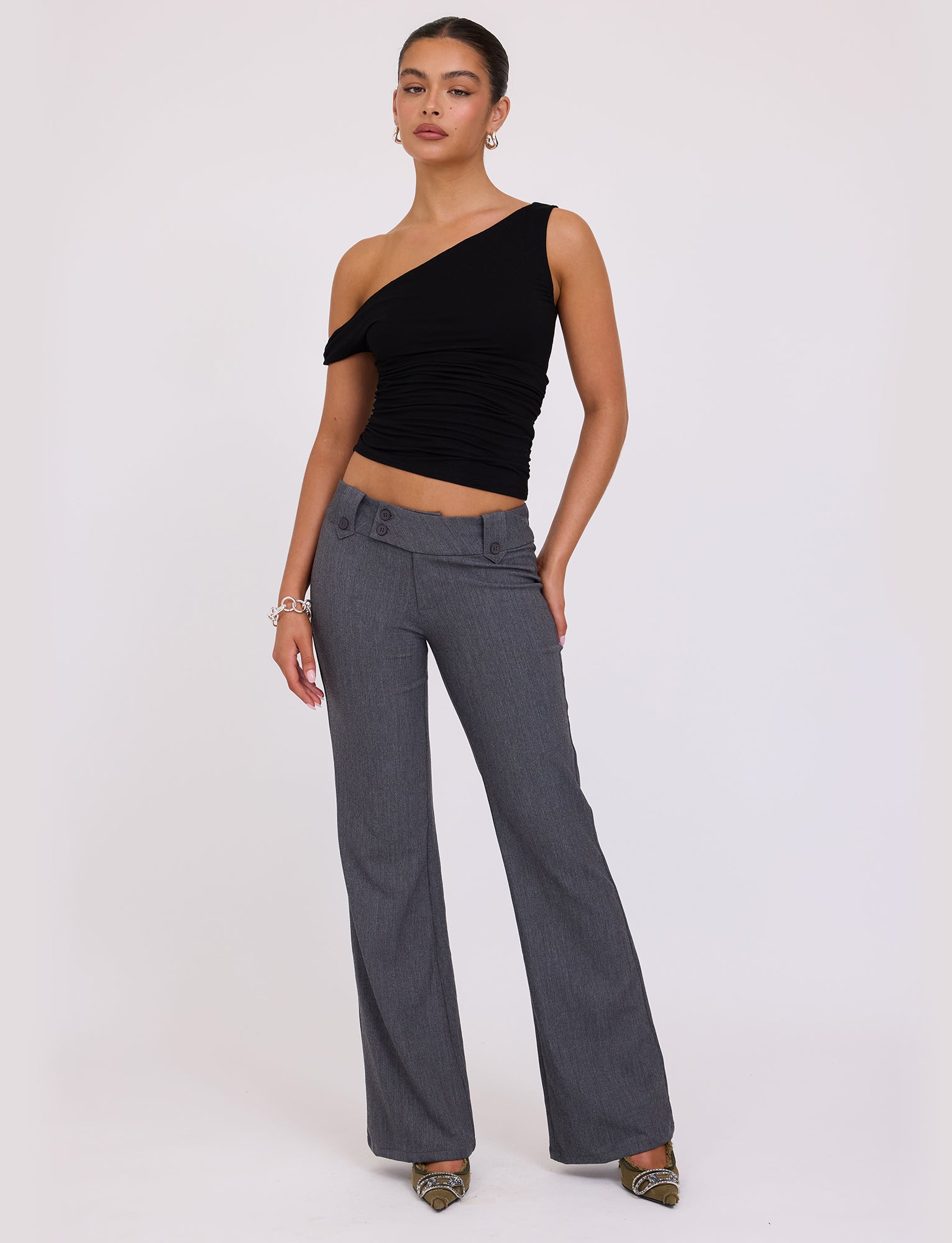 A woman stands against a plain background wearing the KADENCE TOP - BLACK from TIGERMIST and gray high-waisted, wide-leg pants. She has her hair tied back, accessorizes with a bracelet, and wears pointed toe shoes. Her pose is relaxed with one hand resting on her hip.