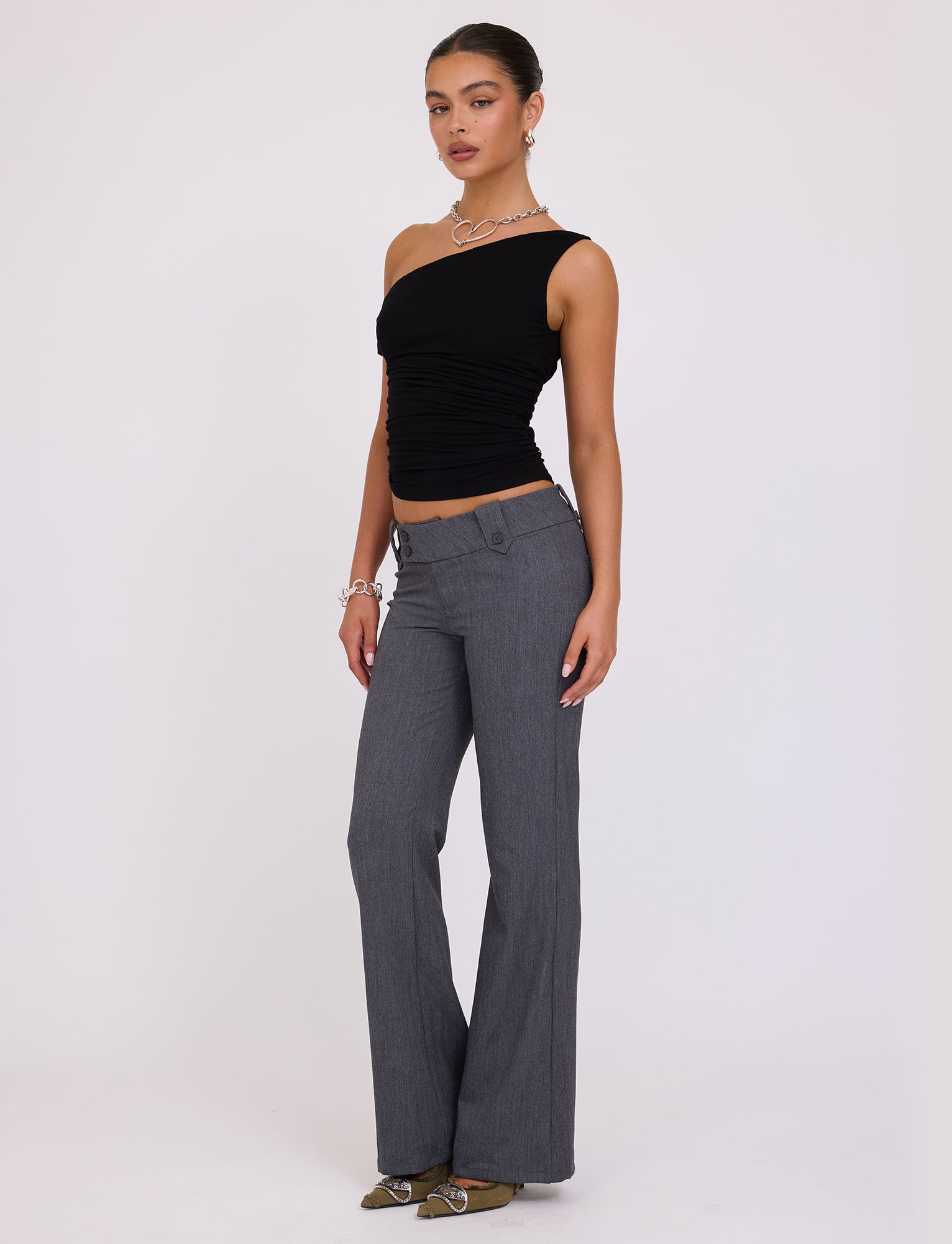 A woman posing against a plain background is wearing the KADENCE TOP - BLACK by TIGERMIST, gray flared pants, and beige pointed shoes. She accessorizes with a silver necklace, bracelet, and hoop earrings, standing confidently with her right hand on her hip.