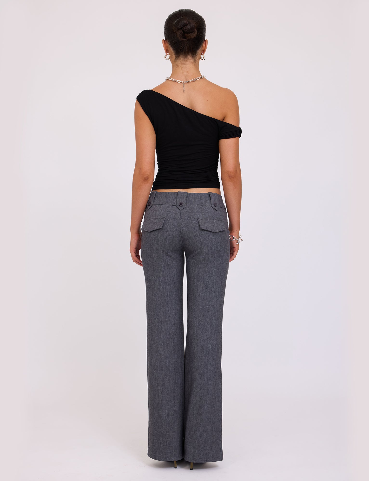 A person with their hair tied up is standing with their back to the camera. They are wearing a black KADENCE TOP from TIGERMIST and gray wide-leg pants. The background is a plain white wall.