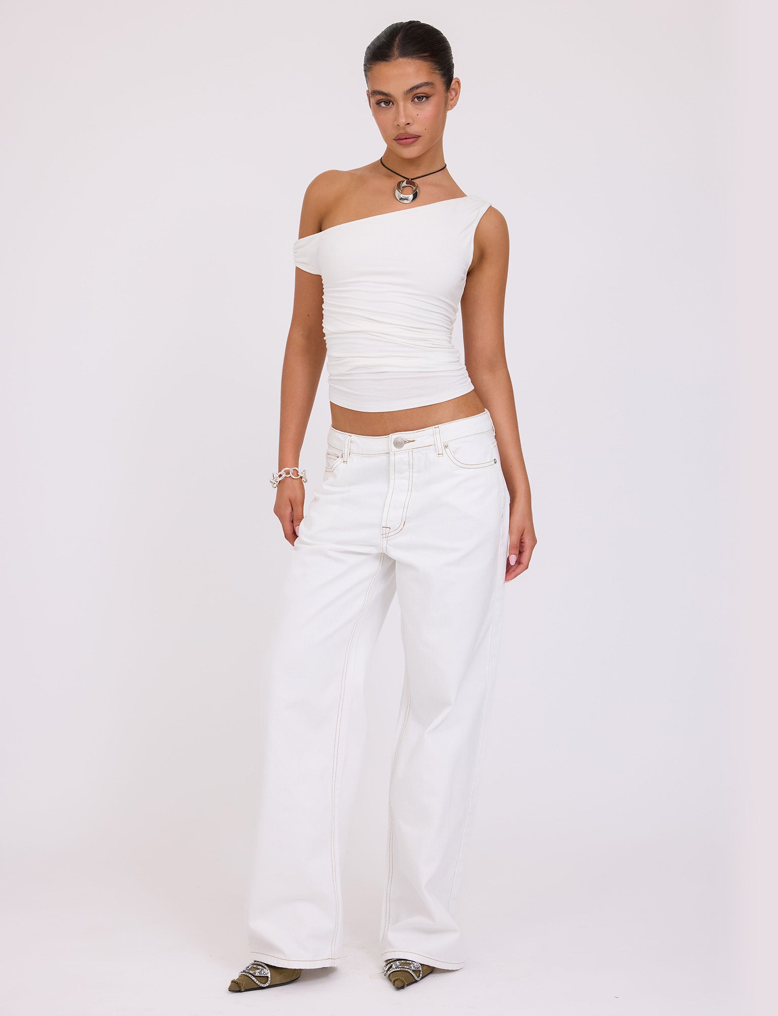 A person with their hair tied back stands against a plain background, donning a chic TIGERMIST KADENCE TOP - WHITE and loose, high-waisted white jeans. They accessorize with a necklace, bracelet, and pointed shoes, striking a relaxed pose.