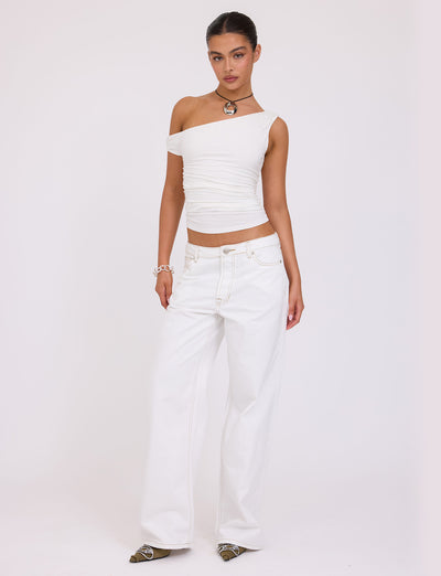 A person with their hair tied back stands against a plain background, donning a chic TIGERMIST KADENCE TOP - WHITE and loose, high-waisted white jeans. They accessorize with a necklace, bracelet, and pointed shoes, striking a relaxed pose.