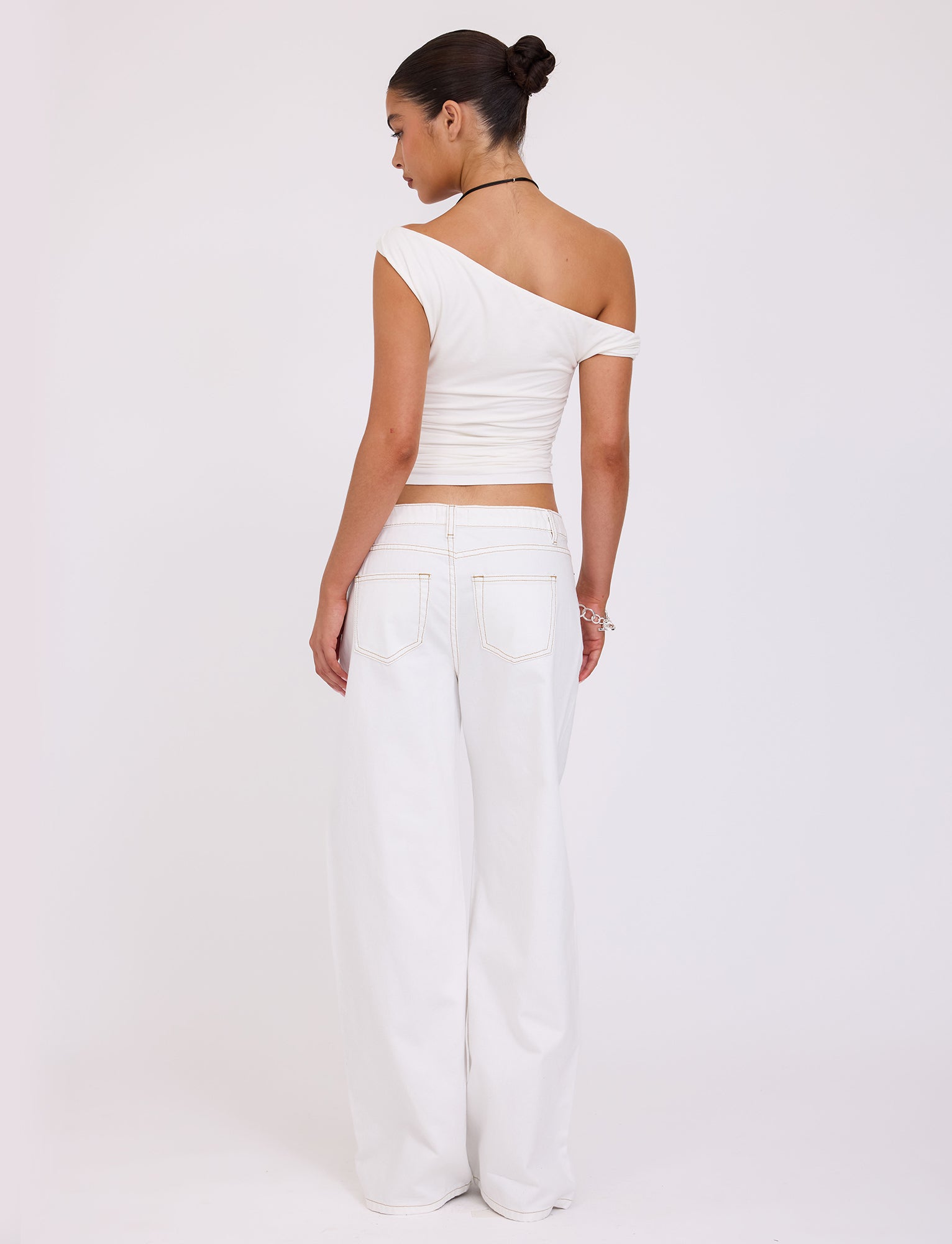 A person with dark hair styled in a bun, wearing the KADENCE TOP - WHITE by TIGERMIST along with high-waisted, wide-leg TIGERMIST white pants, stands with their back to the camera against a plain white background. They are accessorized with bracelets and a necklace.