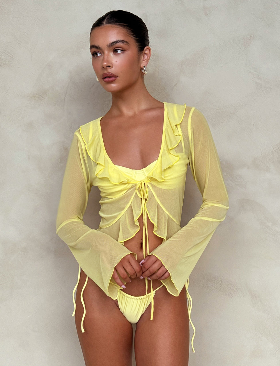 A woman stands against a textured beige background, wearing the TIGERMIST CORVIN TOP - YELLOW : LEMON with a ruched front and ruffle neckline. She pairs it with matching yellow bikini bottoms. Her hair is pulled back, and she wears small hoop earrings.