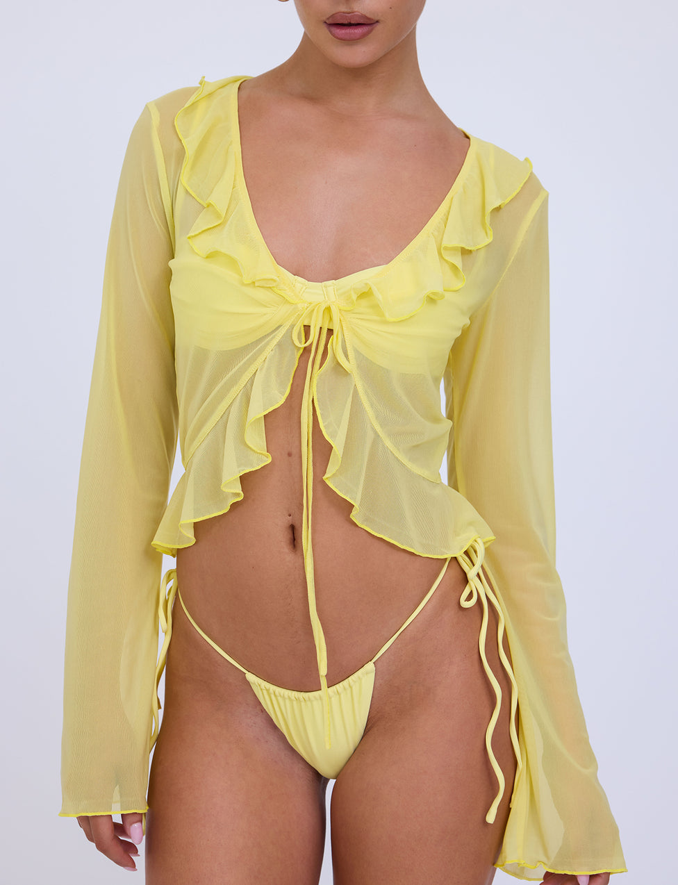 A person is wearing the TIGERMIST CORVIN TOP - YELLOW : LEMON, featuring sheer, long sleeves, ruffled details, and a tie closure in the front. They are also dressed in a matching yellow bikini bottom. The plain white background perfectly highlights the vibrant outfit.