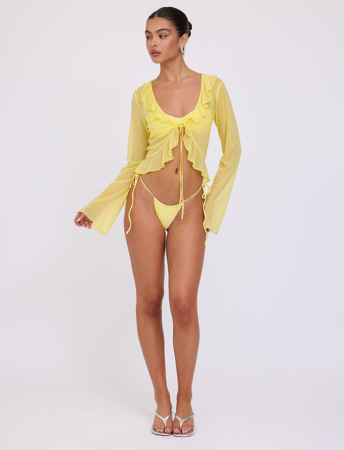 A model stands against a plain white background wearing the TIGERMIST Corvin Top in a vibrant Lemon colourway with long sleeves, a tie-front detail, and ruffled edges. She pairs it with a matching string bikini bottom and clear strap high-heeled sandals. Her hair is styled in a bun, and she gazes to the side.