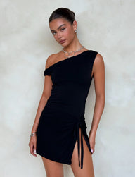 A person with dark hair stands against a light background, wearing the KADENCE DRESS - BLACK by TIGERMIST. The black one-shoulder dress has a knot detail on the side and an asymmetrical neckline, featuring a high split that adds to its elegance. They accessorize with silver hoop earrings, a heart-shaped necklace, and a bracelet on their left wrist.