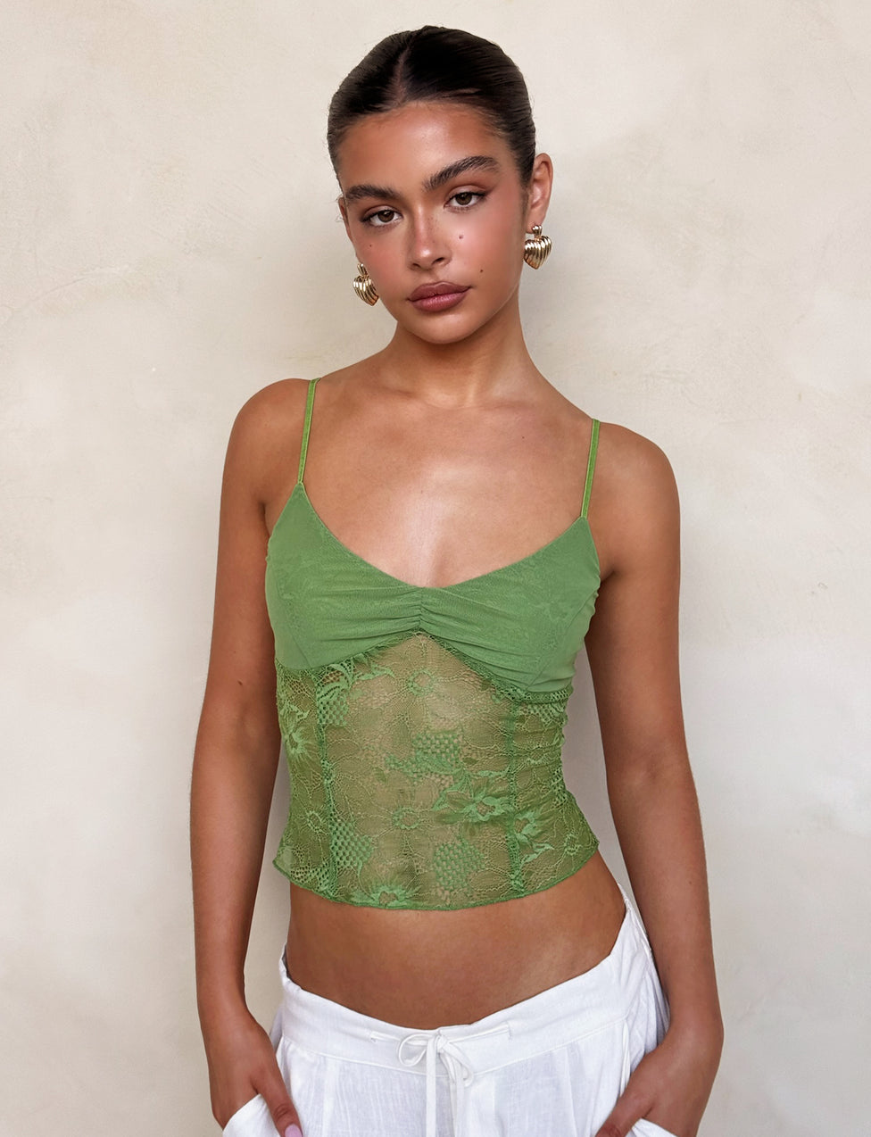 A person stands in front of a textured light beige wall. They wear the SASKIA TOP - GREEN from TIGERMIST paired with white pants. Their hair is pulled back, and they wear hoop earrings. They look directly at the camera with a neutral expression.