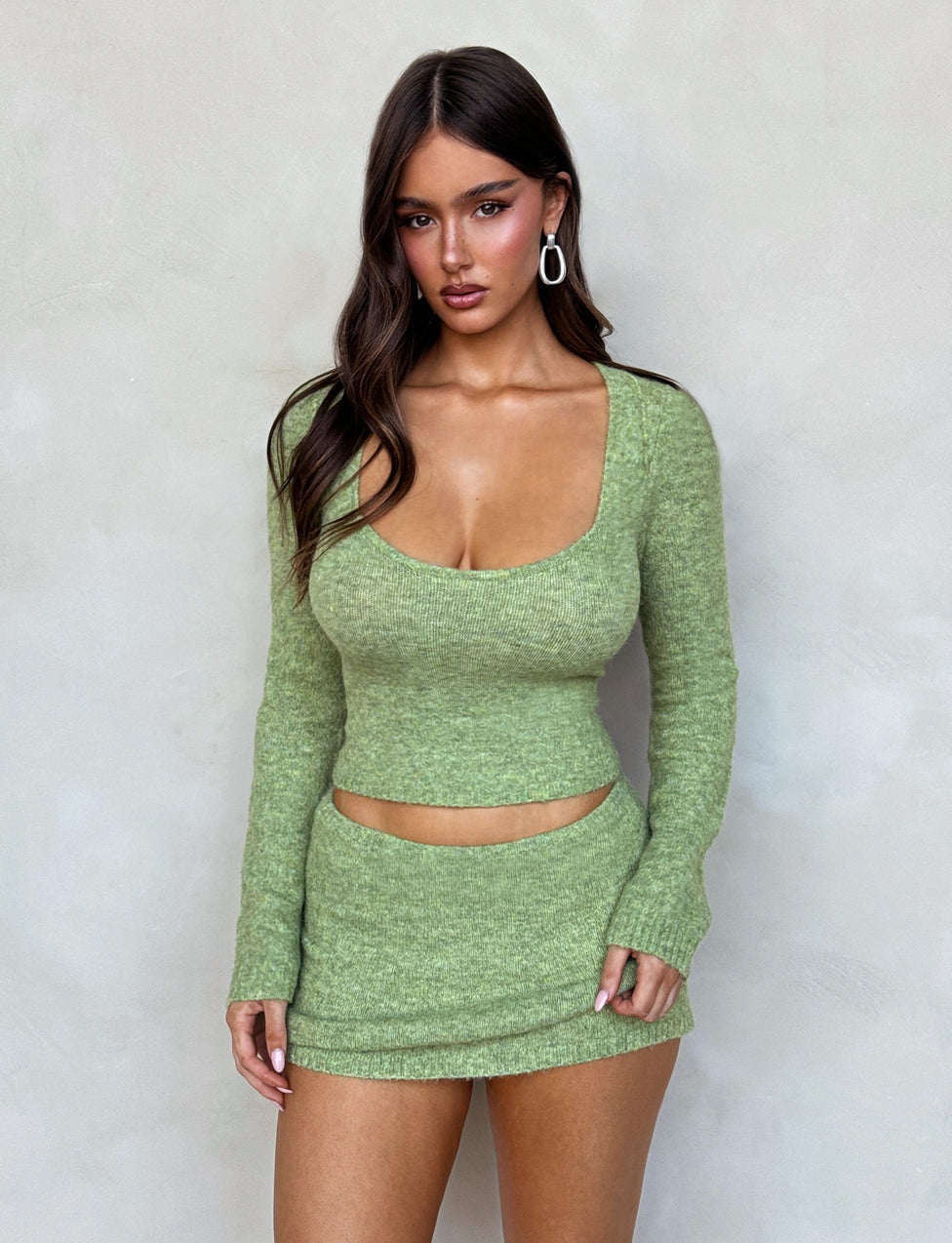 A woman wearing the TIGERMIST LYRICA JUMPER in green, a long-sleeve knit crop top paired with a matching mini skirt. She has long brown hair, hoop earrings, and poses against a light grey background.