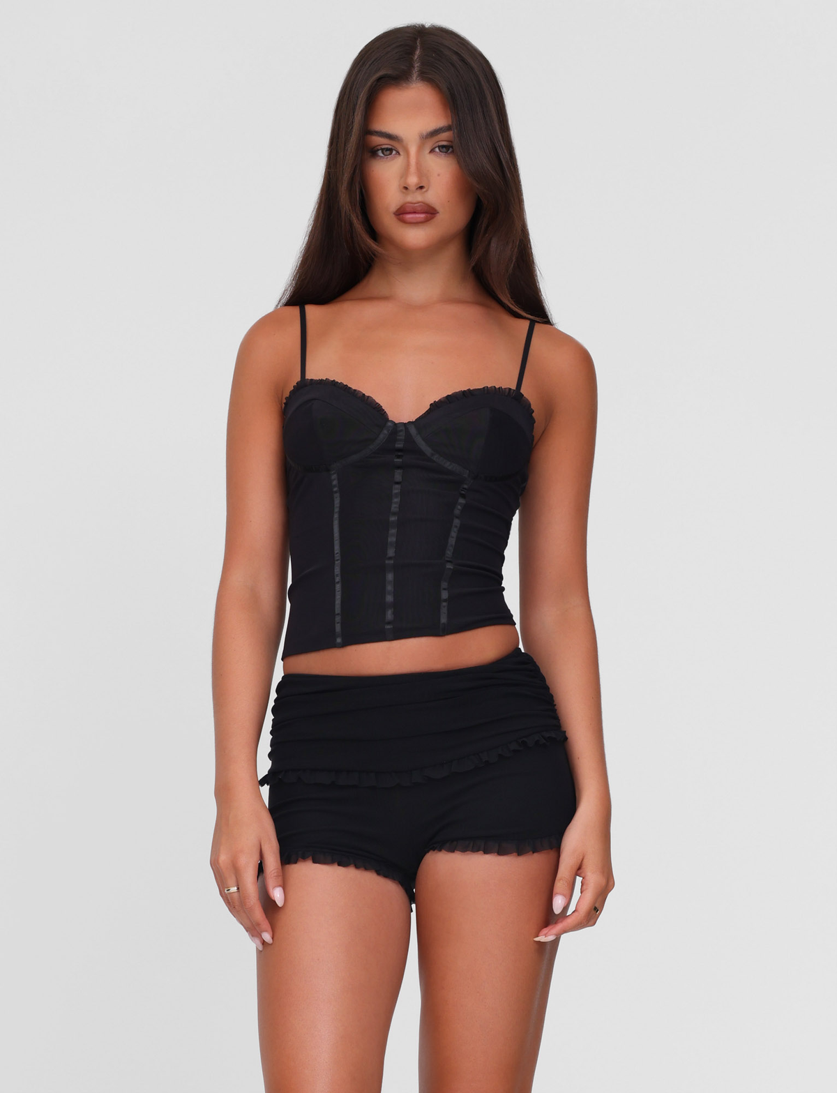 A woman with long dark hair stands against a light gray background, wearing TIGERMIST's GINNY SHORT - BLACK and a fitted black camisole, showcasing the chic bodycon silhouette as she looks directly at the camera with a neutral expression.