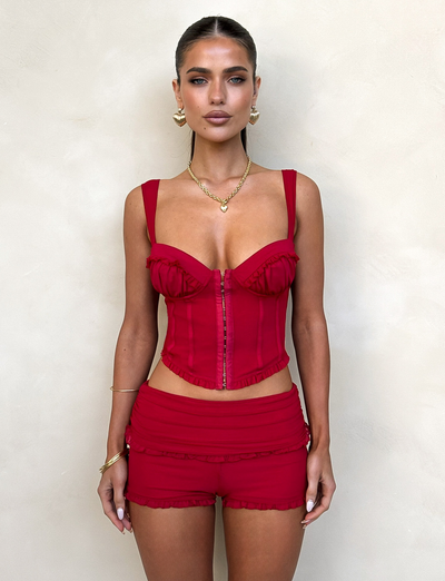 A woman with slicked-back hair poses in TIGERMIST's CHERRY RED GINNY SHORT and matching bustier top, featuring ruffle details and gold jewelry, set against a neutral light background.