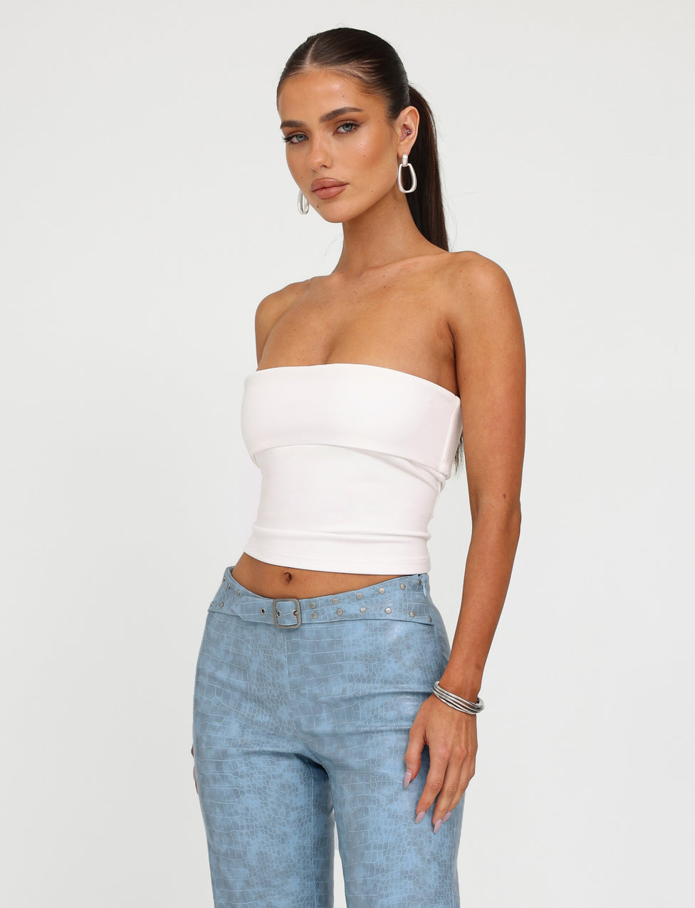 A woman with long, dark hair tied back in a sleek ponytail is wearing the DARIELLE TOP in white from TIGERMIST along with light blue, high-waisted pants. She accessorizes with hoop earrings and several bracelets on her right wrist. The background is plain white.