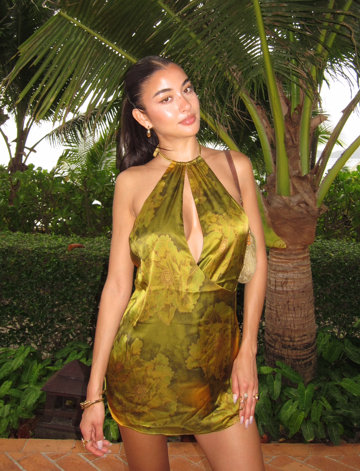 A woman poses outdoors in front of palm trees, wearing the TIGERMIST MONROE DRESS - GREEN: WATER COLOUR FLORAL with a halter neckline. She styles her long dark hair back, accessorizes with gold jewelry, and carries a neutral purse.