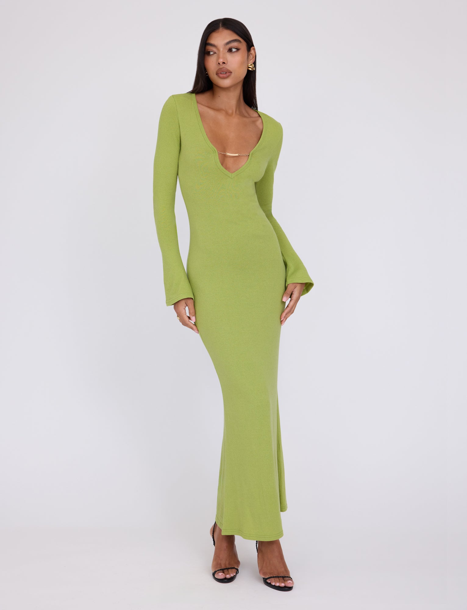A woman with long, dark hair poses in the light green, long-sleeved, form-fitting ROMIE DRESS - GREEN : MATCHA GREEN by TIGERMIST. The dress features a deep V-neckline with a delicate strap detail. She pairs it with black high-heeled sandals and stands against a plain white background.