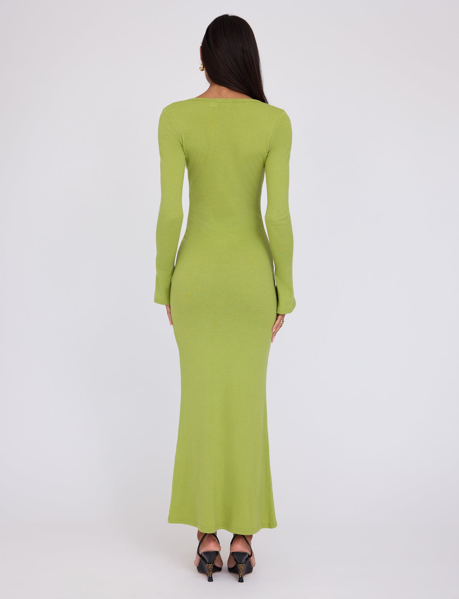 A person with long dark hair stands facing away, wearing the floor-length, fitted ROMIE DRESS in Matcha Green from TIGERMIST. The individual is also wearing black high-heeled shoes. The background is plain white, drawing attention to the clothes.