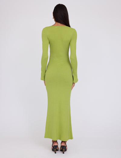 A person with long dark hair stands facing away, wearing the floor-length, fitted ROMIE DRESS in Matcha Green from TIGERMIST. The individual is also wearing black high-heeled shoes. The background is plain white, drawing attention to the clothes.