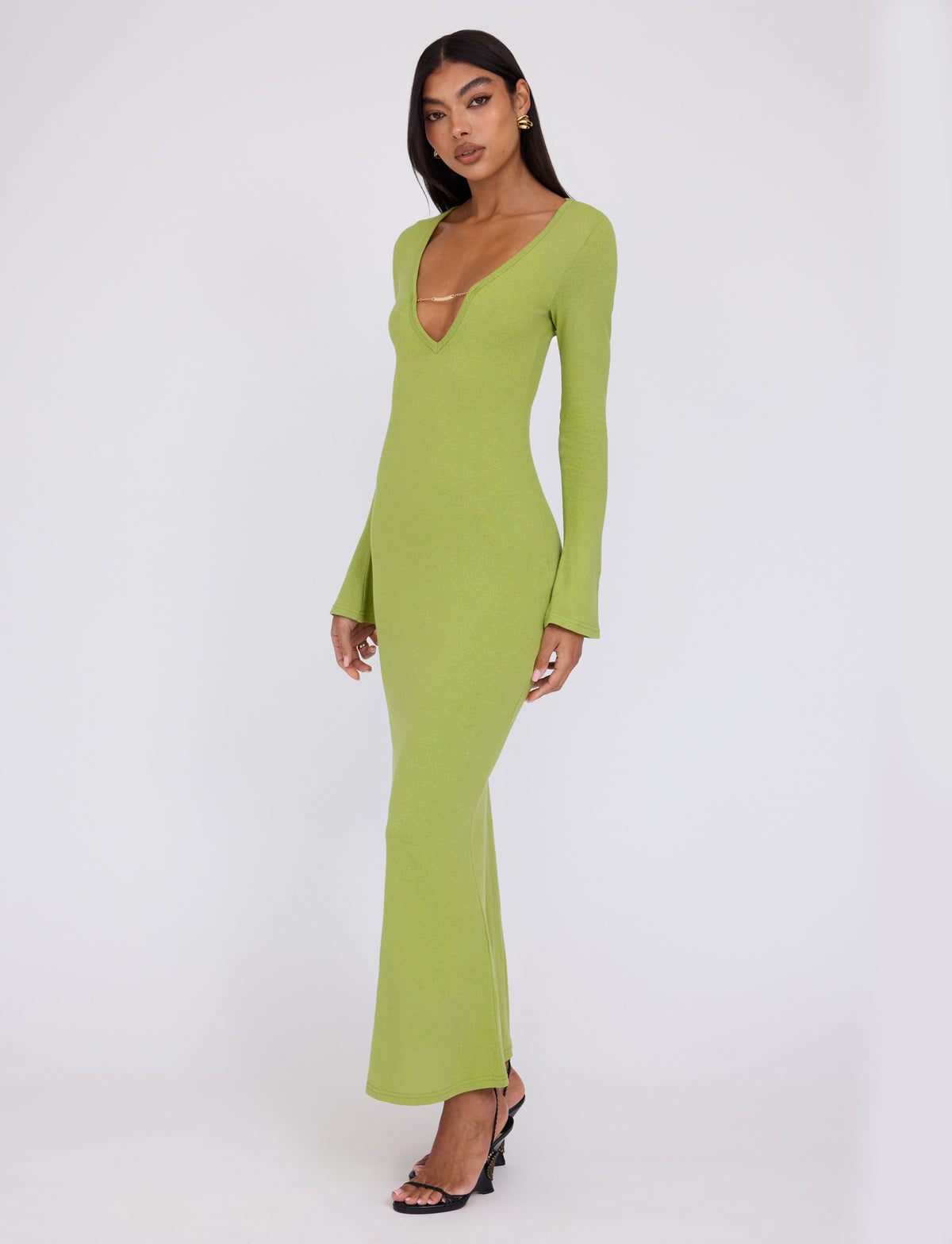 A person stands against a plain white background, wearing the long-sleeved, floor-length ROMIE DRESS - GREEN : MATCHA GREEN by TIGERMIST with a deep V-neck. They have long, dark hair and are wearing black open-toed heels. Their hands rest at their sides, and they have a neutral expression.