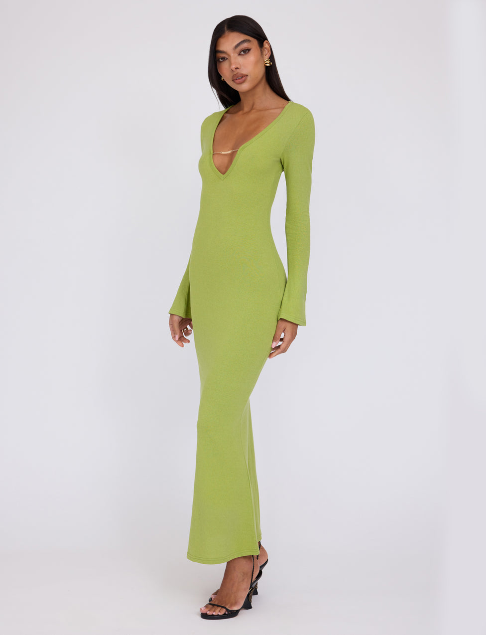 A person stands against a plain white background, wearing the long-sleeved, floor-length ROMIE DRESS - GREEN : MATCHA GREEN by TIGERMIST with a deep V-neck. They have long, dark hair and are wearing black open-toed heels. Their hands rest at their sides, and they have a neutral expression.