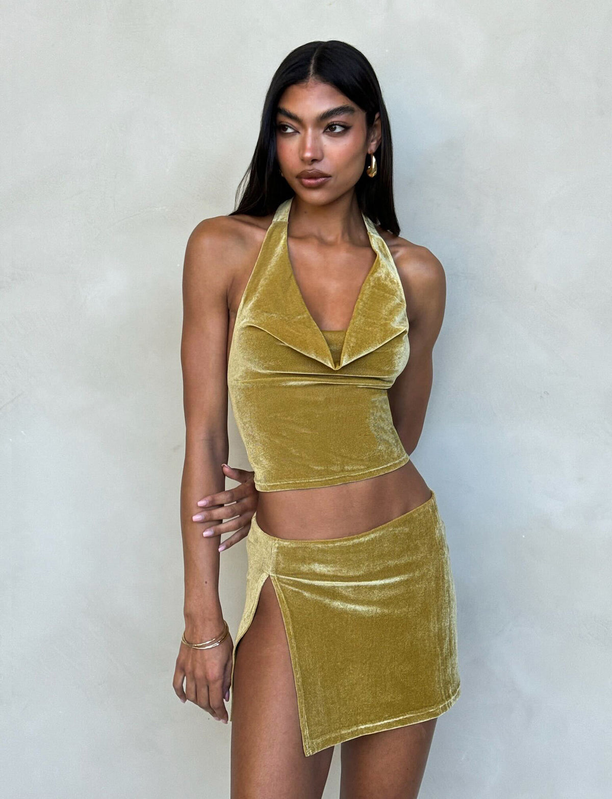 A woman with long dark hair models a shiny, gold velvet two-piece outfit from TIGERMIST against a plain light-gray background. She wears the HARLOWE TOP, featuring an open draped neckline, along with a matching mini skirt that has a high slit on the left side. Gold hoop earrings complete her look.