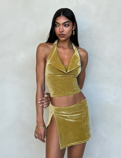 A woman with long dark hair models a shiny, gold velvet two-piece outfit from TIGERMIST against a plain light-gray background. She wears the HARLOWE TOP, featuring an open draped neckline, along with a matching mini skirt that has a high slit on the left side. Gold hoop earrings complete her look.