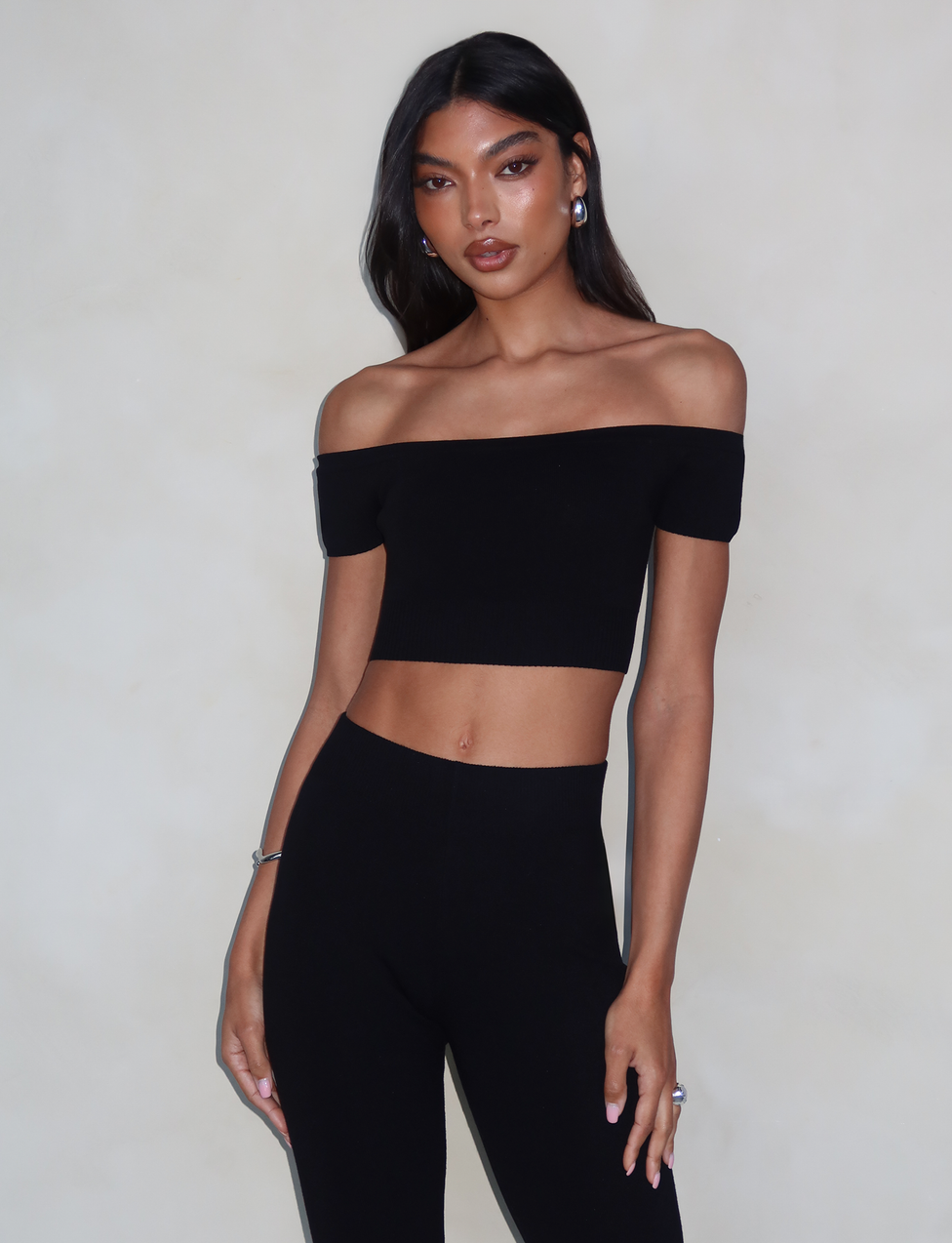 A person with long, dark hair dons the chic LEDGER TOP - BLACK by TIGERMIST paired with high-waisted black pants, creating a sleek silhouette. They stand against a neutral background, with one arm relaxed at their side and the other slightly bent, showcasing a confident pose.