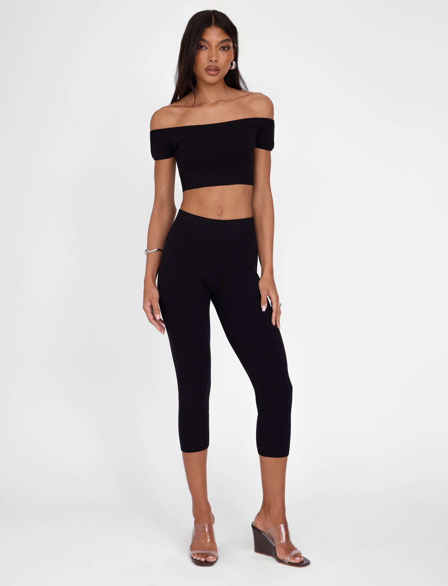 A woman stands against a plain white background, wearing a black off-the-shoulder crop top and TIGERMIST's LEDGER CAPRI in black. She has long dark hair and is sporting clear, open-toe wedge sandals. Her left hand rests by her side while her right hand is confidently on her hip.