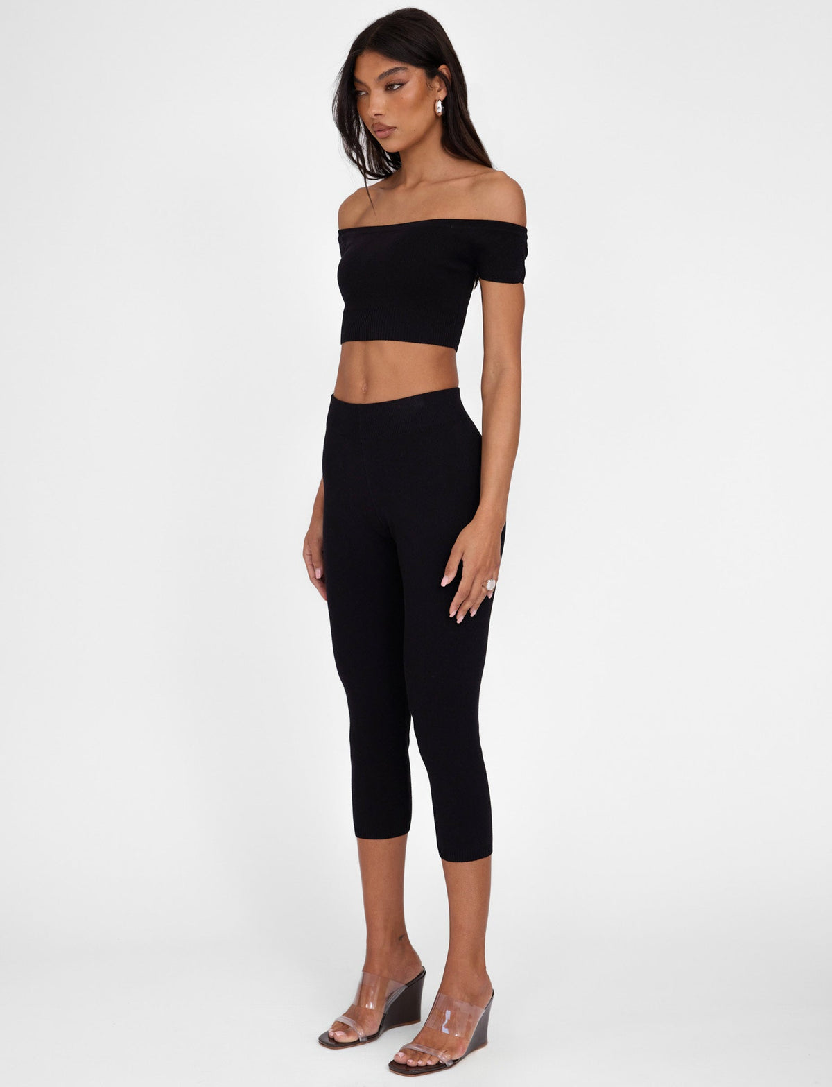 A woman stands against a plain white background, wearing a black off-the-shoulder crop top paired with TIGERMIST's LEDGER CAPRI in black. She is also adorned with clear open-toe wedge sandals and hoop earrings. Her long, dark hair flows as she gazes downward.