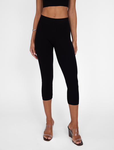 A person wearing a black, sleeveless crop top paired with TIGERMIST's LEDGER CAPRI - BLACK high-rise capri leggings. Their hands rest by their sides, and they are sporting clear wedge-heeled sandals against a plain white background.