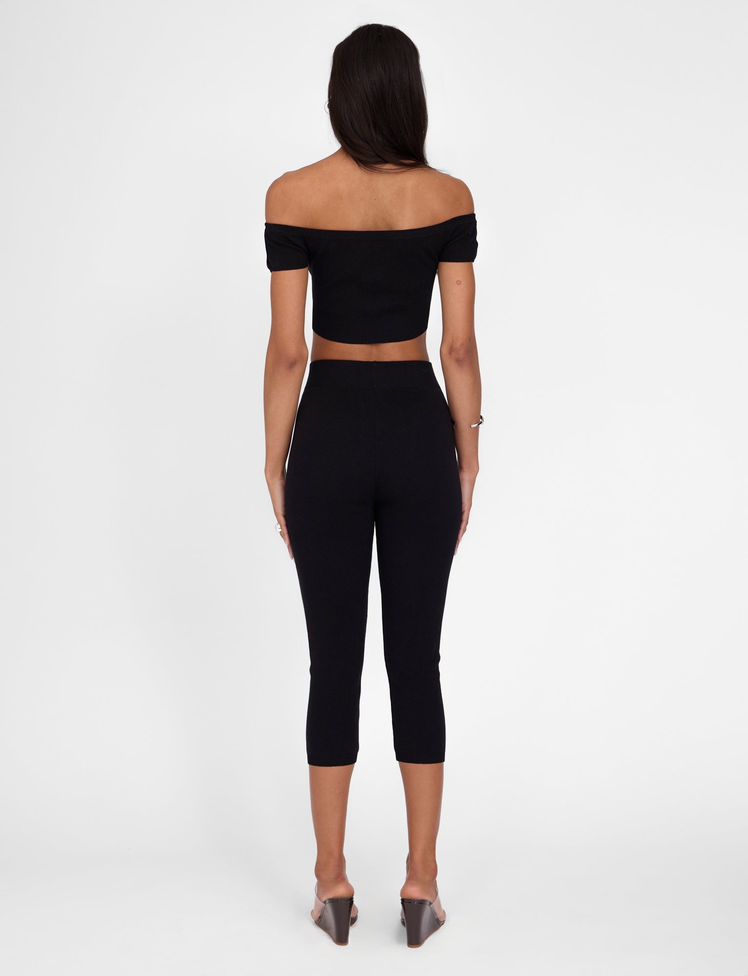 A woman with long dark hair stands with her back facing the camera, wearing an off-the-shoulder black crop top and TIGERMIST's LEDGER CAPRI in black. The high-rise waist capri pants have a bodycon fit, and she's also sporting transparent high-heeled shoes. The background is white and uncluttered.