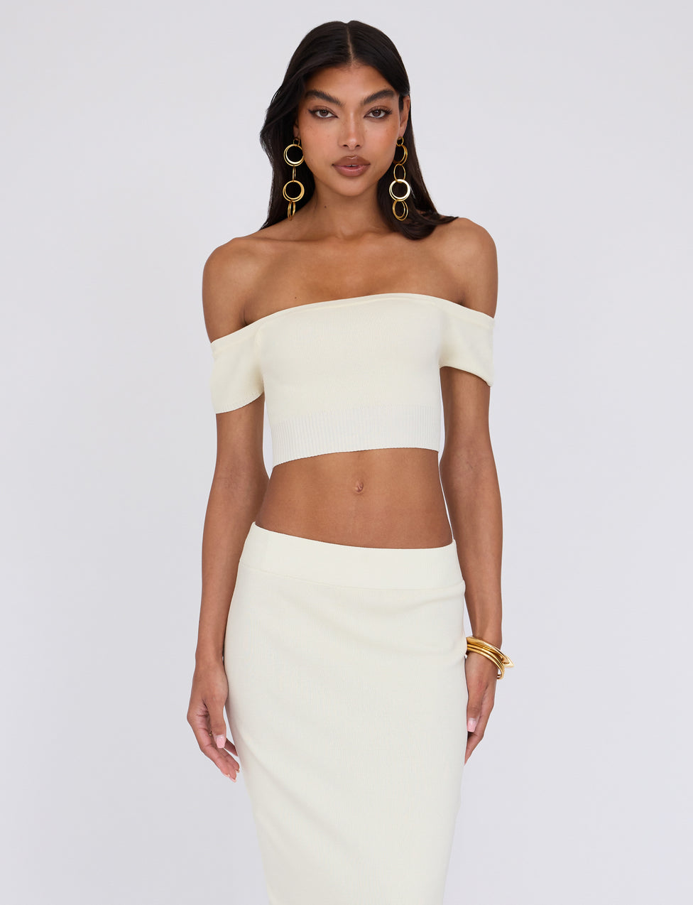 A woman with long dark hair stands against a plain background, wearing the cream LEDGER TOP - WHITE from TIGERMIST and a matching high-waisted skirt. She has bold hoop earrings and gold bangles on her left wrist, looking confidently at the camera with a relaxed posture.