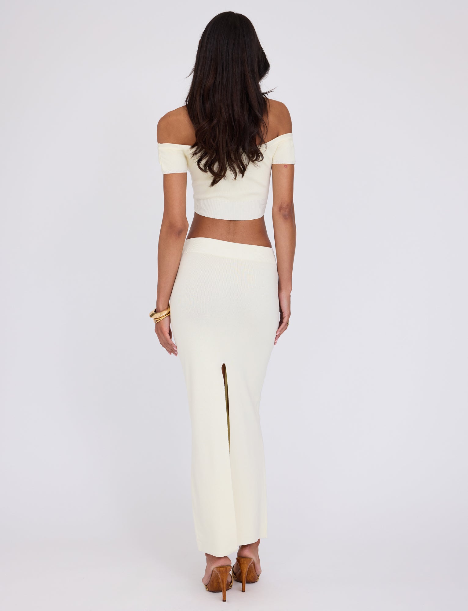 A woman with long, dark hair is seen from the back, standing against a plain white background. She is wearing an off-the-shoulder, cropped, short-sleeve top and the TIGERMIST LEDGER SKIRT - WHITE : CREAM, a high-waisted, ankle-length skirt with a high slit at the back. Her chic ensemble epitomizes TIGERMIST's style as she stands confidently in high heels.