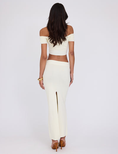A woman with long, dark hair is seen from the back, standing against a plain white background. She is wearing an off-the-shoulder, cropped, short-sleeve top and the TIGERMIST LEDGER SKIRT - WHITE : CREAM, a high-waisted, ankle-length skirt with a high slit at the back. Her chic ensemble epitomizes TIGERMIST's style as she stands confidently in high heels.