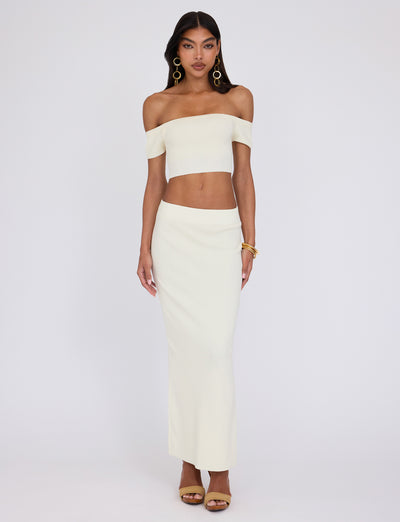 A woman is posing against a plain background, wearing an off-the-shoulder, cream-colored crop top and the high-waisted LEDGER SKIRT - WHITE : CREAM from TIGERMIST. She accessorizes with large hoop earrings, gold bangles, and golden high-heeled sandals and has long, dark hair.