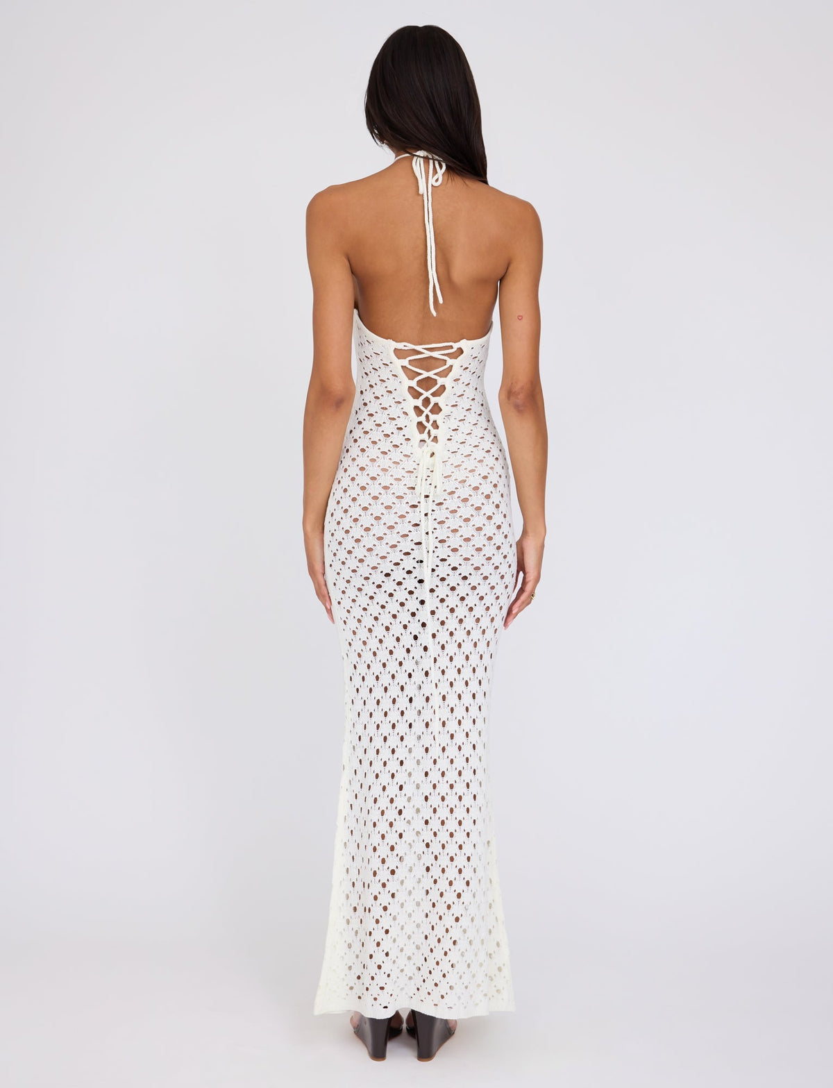 A woman stands with her back to the camera, showcasing the CIARAN DRESS - WHITE by TIGERMIST. The white crocheted, halter-neck dress has an open back adorned with intricate patterns and ties. Her long, dark hair cascades down her back as she poses against a plain white background.