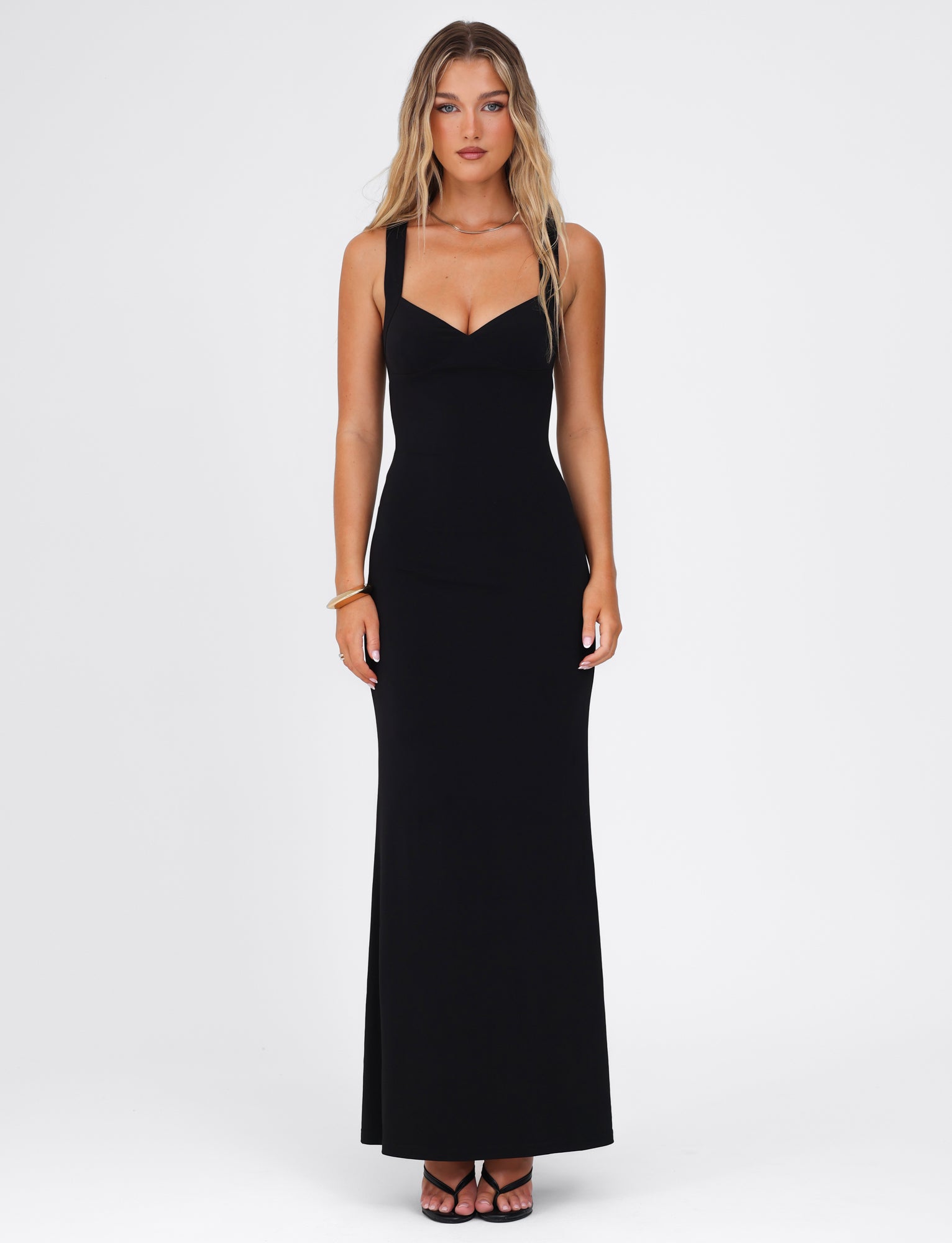A woman stands against a plain white background, modeling the TIGERMIST Sonnet Maxi Dress in black, which showcases a fitted silhouette with a sweetheart neckline and an elegant open back design. She has long, wavy hair and wears black open-toed shoes, maintaining a neutral expression while her right wrist is adorned with a bracelet.