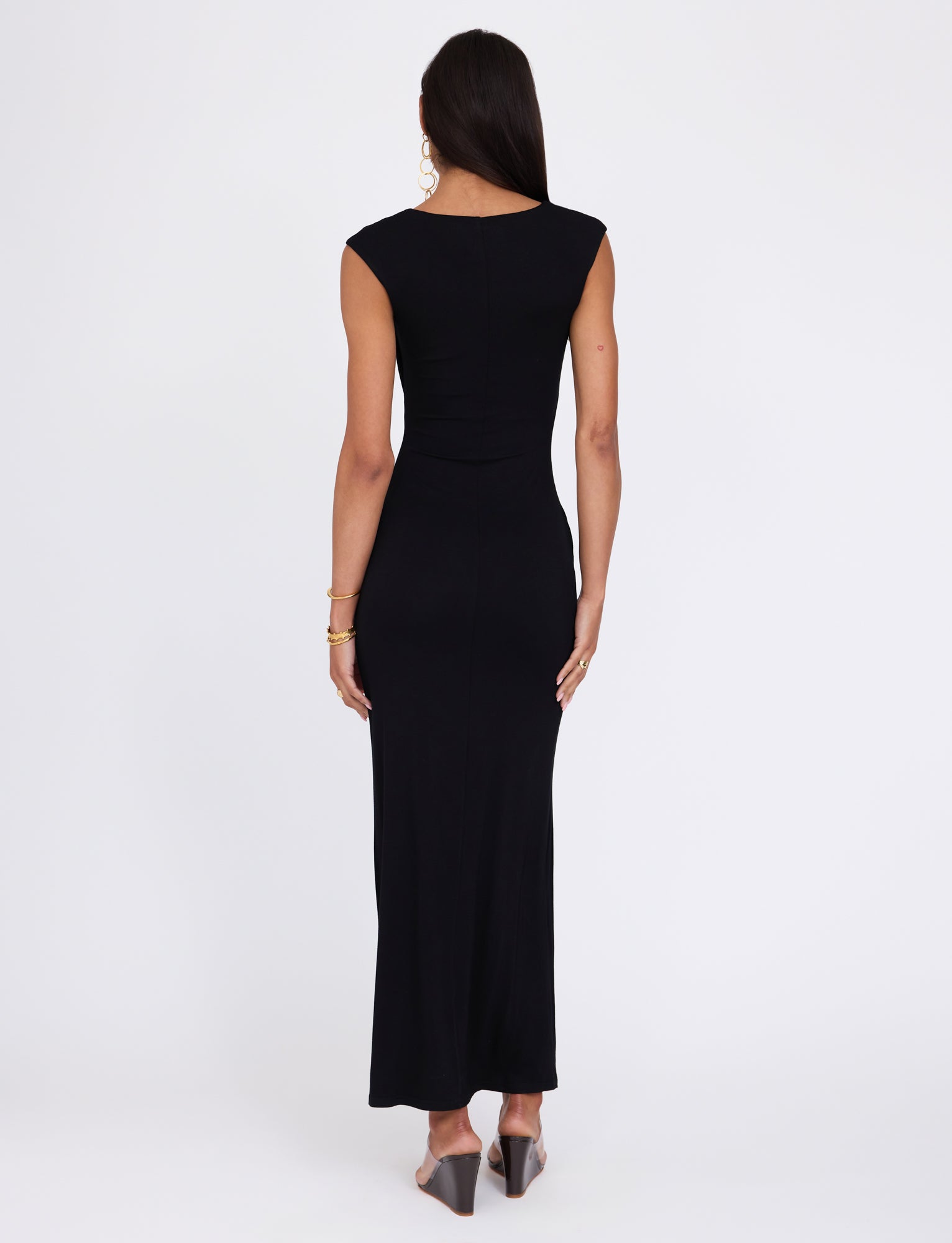 A woman with long brown hair is standing with her back to the camera, wearing the fitted, sleeveless BLAKELY DRESS - BLACK by TIGERMIST that reaches her ankles. She is accessorized with gold hoop earrings, gold bracelets, and black heeled mules. The background is plain white.