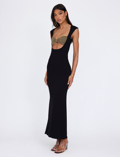 A woman with long hair is wearing the Blakely Dress in black by TigerMist, featuring a plunging neckline that reveals a tan bandeau underneath. She accessorizes with gold hoop earrings, bracelets, and clear high-heeled sandals while standing against a plain white background.