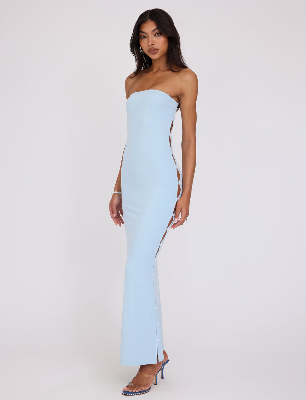 A woman stands confidently in the TIGERMIST ROMINA DRESS - BLUE, a strapless gown featuring side cutouts held together by small buttons. She accessorizes with a bracelet and high-heeled sandals, striking a pose on a plain white background.