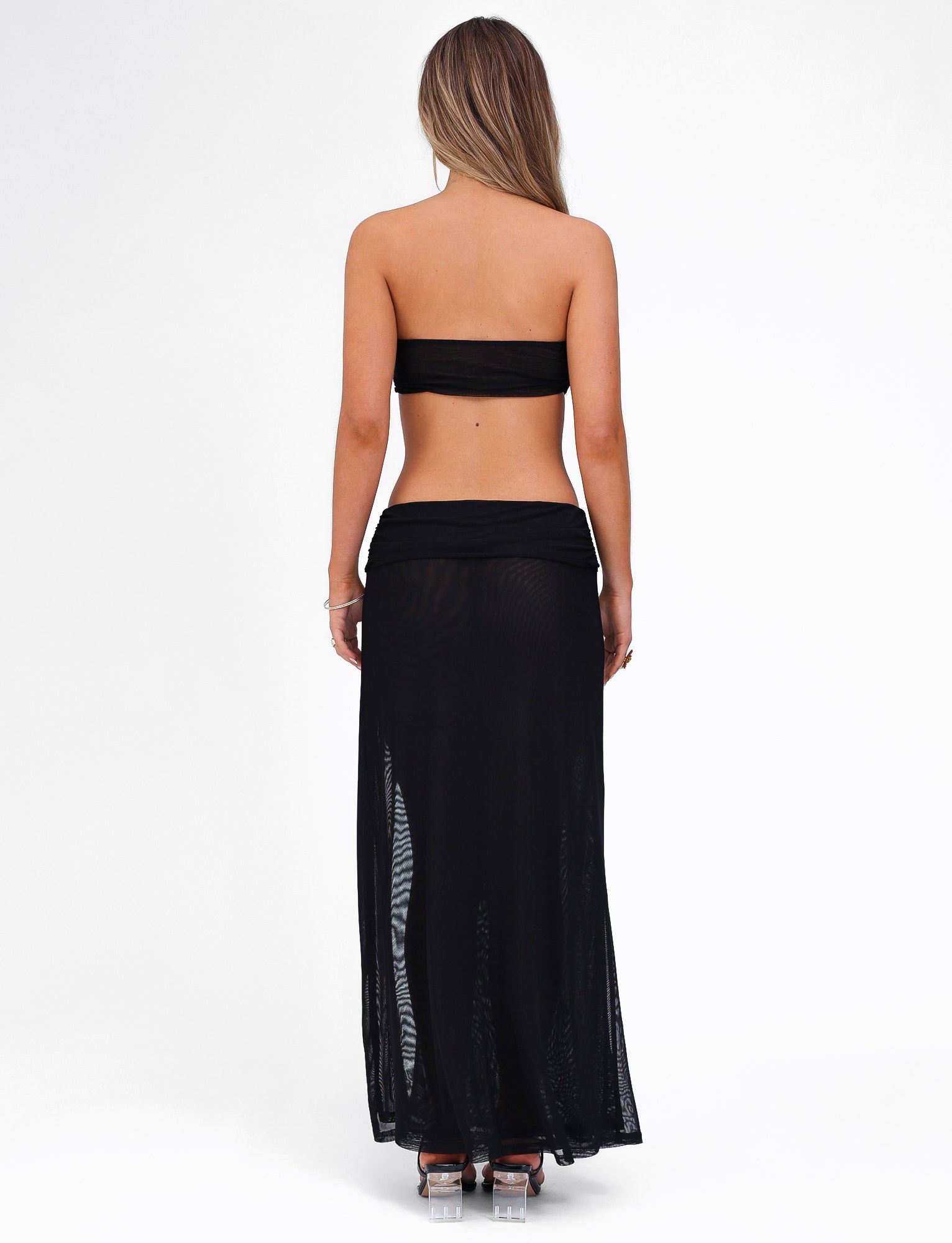 A woman is standing with her back to the camera, wearing the black KYNLEE MAXI DRESS by TIGERMIST. She has long, light brown hair and is paired the dress with high-heeled shoes. The background is plain white.