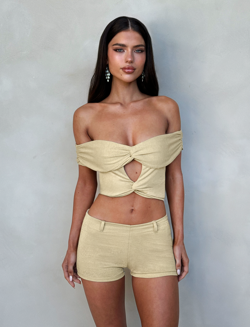 A woman with long, dark hair stands against a plain light-colored background. She is wearing the stylish TIGERMIST LYNSEY TOP in gold, featuring an off-the-shoulder design with a front twist and keyhole cutout, paired with matching high-waisted shorts. She also has on dangle earrings.
