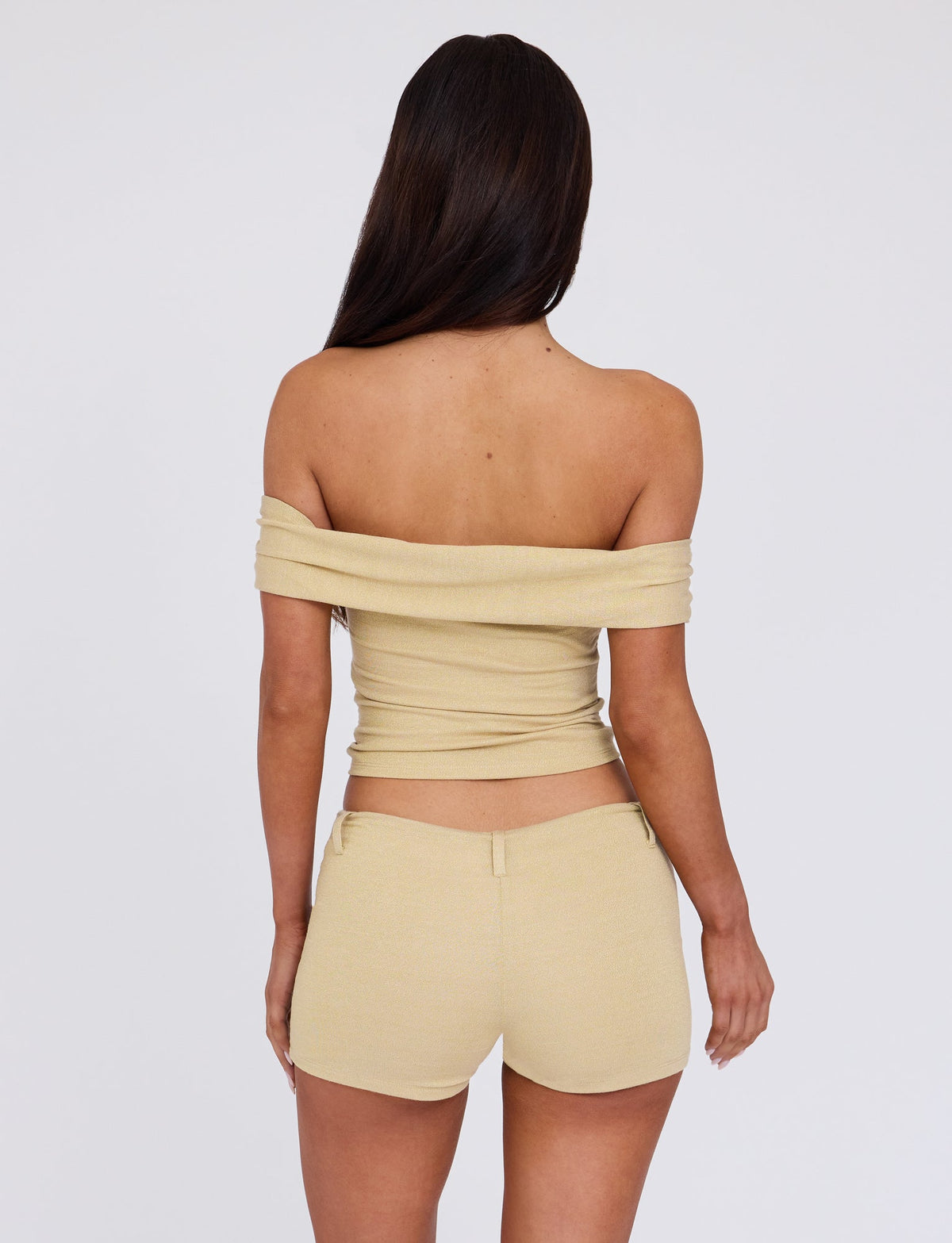 A person with long, straight hair stands facing away from the camera, wearing an off-the-shoulder gold top from Tiger Mist's LYNSEY TOP - GOLD collection and matching beige shorts. The background is plain white.