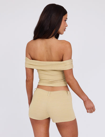 A woman with long dark hair faces away, wearing a beige off-the-shoulder top and matching high-waisted LYNSEY SHORT - GOLD from TIGERMIST. The background is plain white, emphasizing her outfit. She appears to be standing in a relaxed pose with her right arm slightly bent.