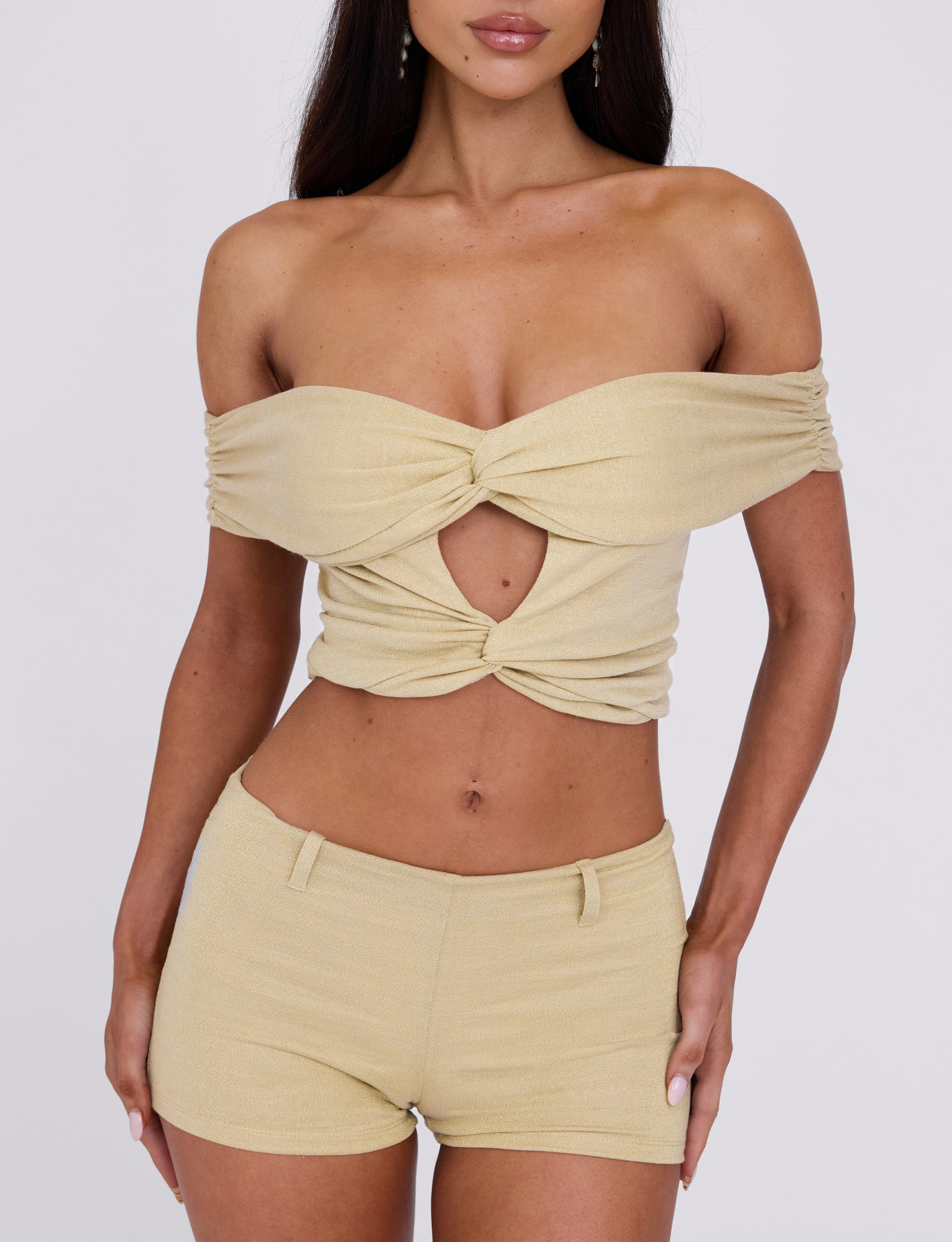 A woman is modeling the LYNSEY SHORT - GOLD, an off-the-shoulder crop top featuring a central twist and keyhole cutout paired with matching high-waisted shorts from TIGERMIST. The outfit is stylish and form-fitting, boasting a minimalistic and chic design against a plain white background.