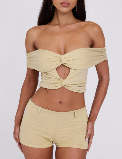 A woman is modeling the LYNSEY SHORT - GOLD, an off-the-shoulder crop top featuring a central twist and keyhole cutout paired with matching high-waisted shorts from TIGERMIST. The outfit is stylish and form-fitting, boasting a minimalistic and chic design against a plain white background.