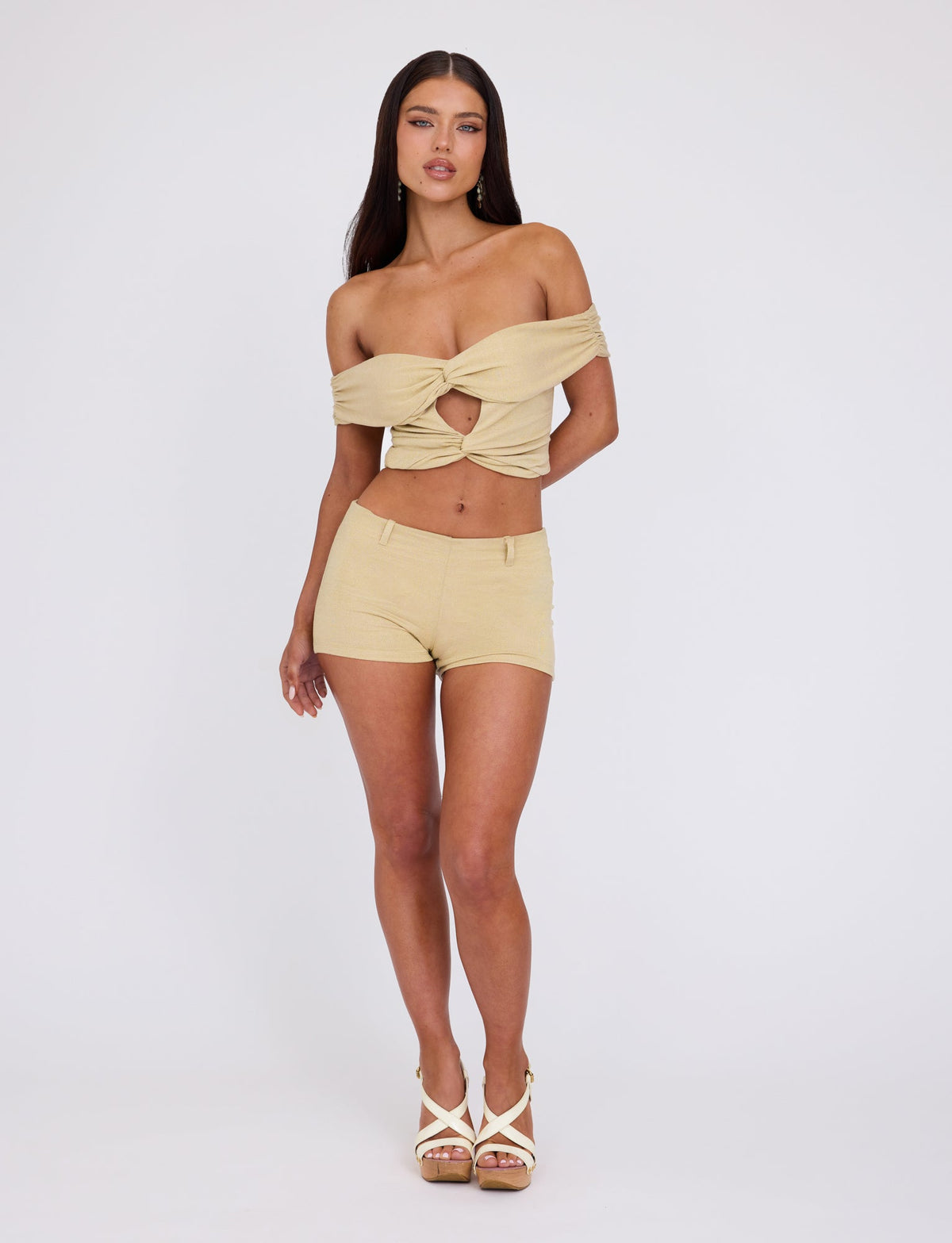 A woman stands against a plain background, wearing the TIGERMIST LYNSEY TOP in gold, an off-shoulder crop top with a front twist and cutout, paired with matching high-waisted shorts. She completes her look with white high-heeled sandals. Her hair is long and straight, and she exudes confidence in her pose.
